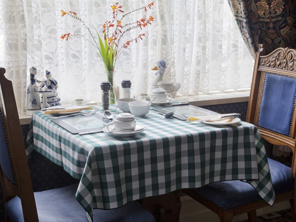 Ruokapöytä on katettu ruutukuosisella pöytäliinalla, sen ympärillä on tuoleja ja pöydällä on maljakko täynnä kukkia.