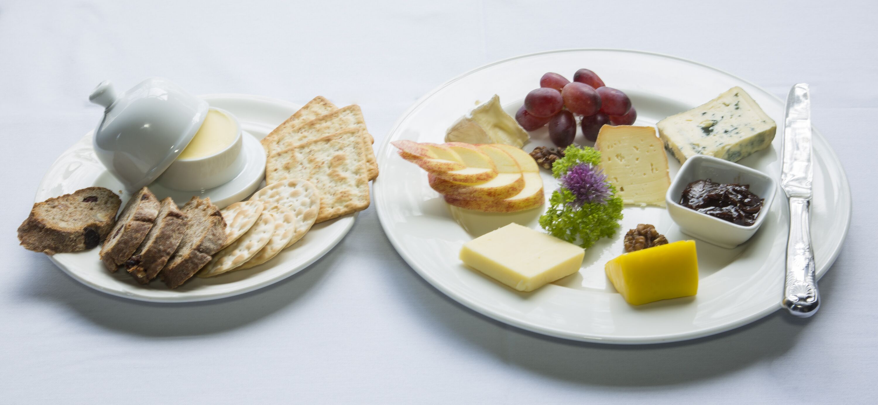 Two plates of cheese, crackers, and fruits on a white table.