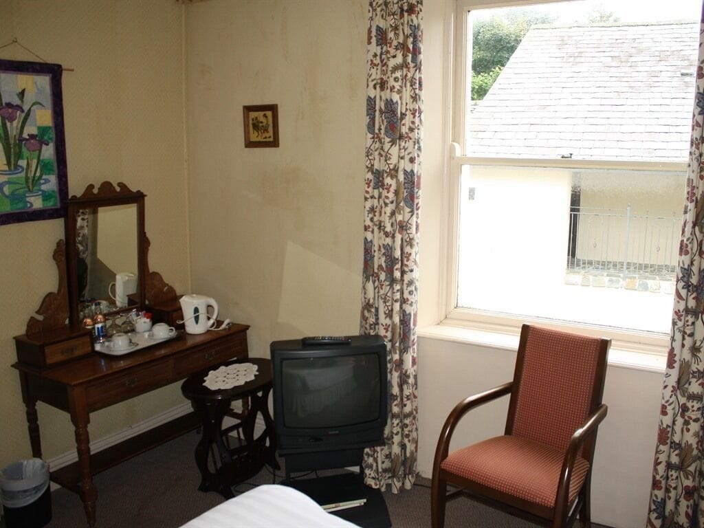 Ein Hotelzimmer mit einem Holzkoffer, einem Fernseher, einem Sessel und einem fenster mit Vorhängen.