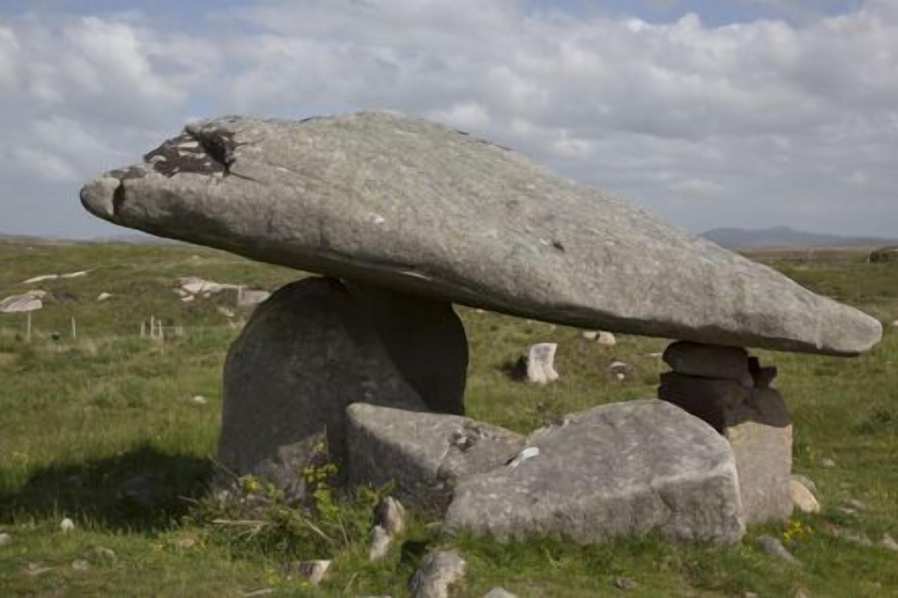 Una estructura de piedra vertical, con una gran losa plana equilibrada sobre una base de piedra más pequeña, rodeada de hierba y otras rocas en un paisaje natural.