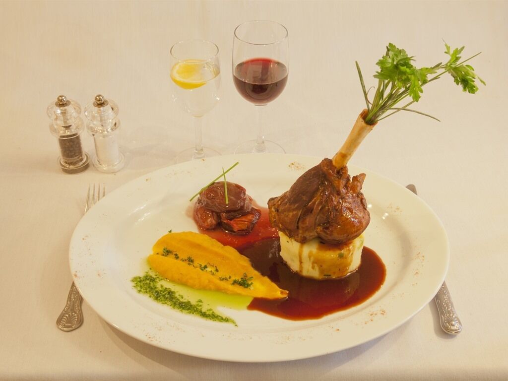 Un plato de comida con un rabo de cordero, verduras y salsa. Hay dos copas de vino y saleros y pimenteros sobre la mesa.