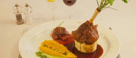 Un plato de comida con un rabo de cordero, verduras y salsa. Hay dos copas de vino y saleros y pimenteros sobre la mesa.