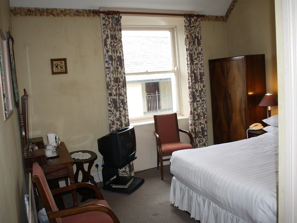 A hotel room with a bed, a television, a chair, a small table, a window with curtains, and a wooden wardrobe.