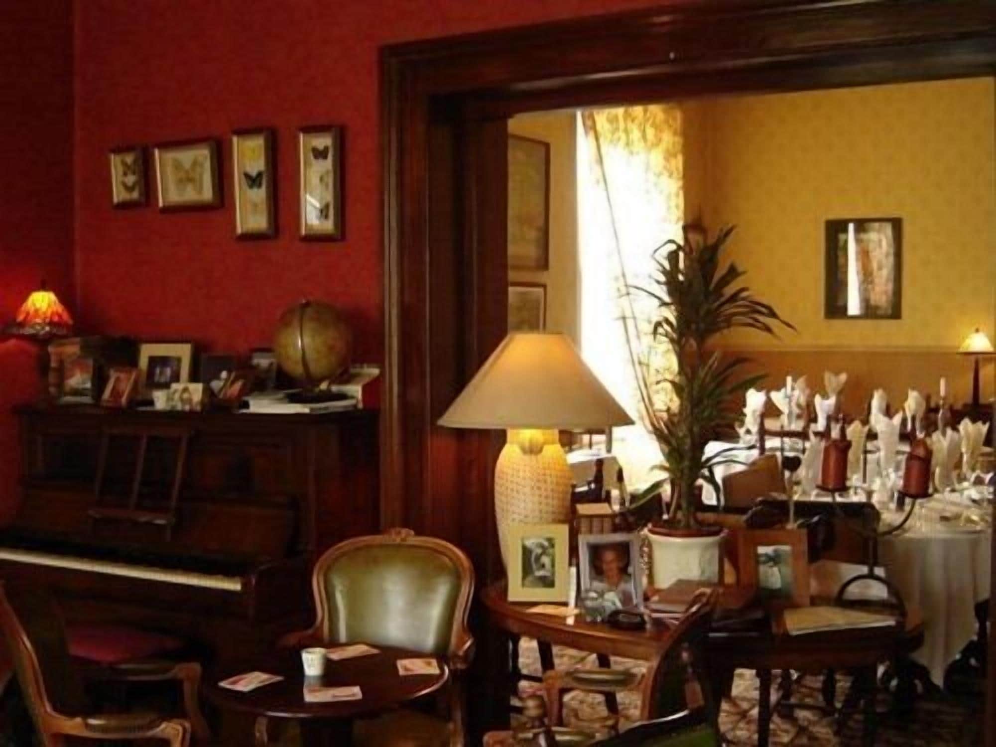 A room with a piano, a table with a lamp, chairs, and framed pictures on the wall.