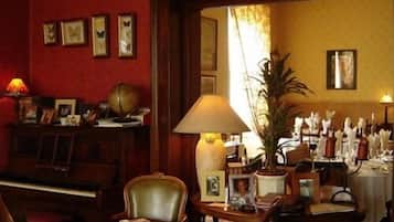 A room with a piano, a table with a lamp, chairs, and framed pictures on the wall.