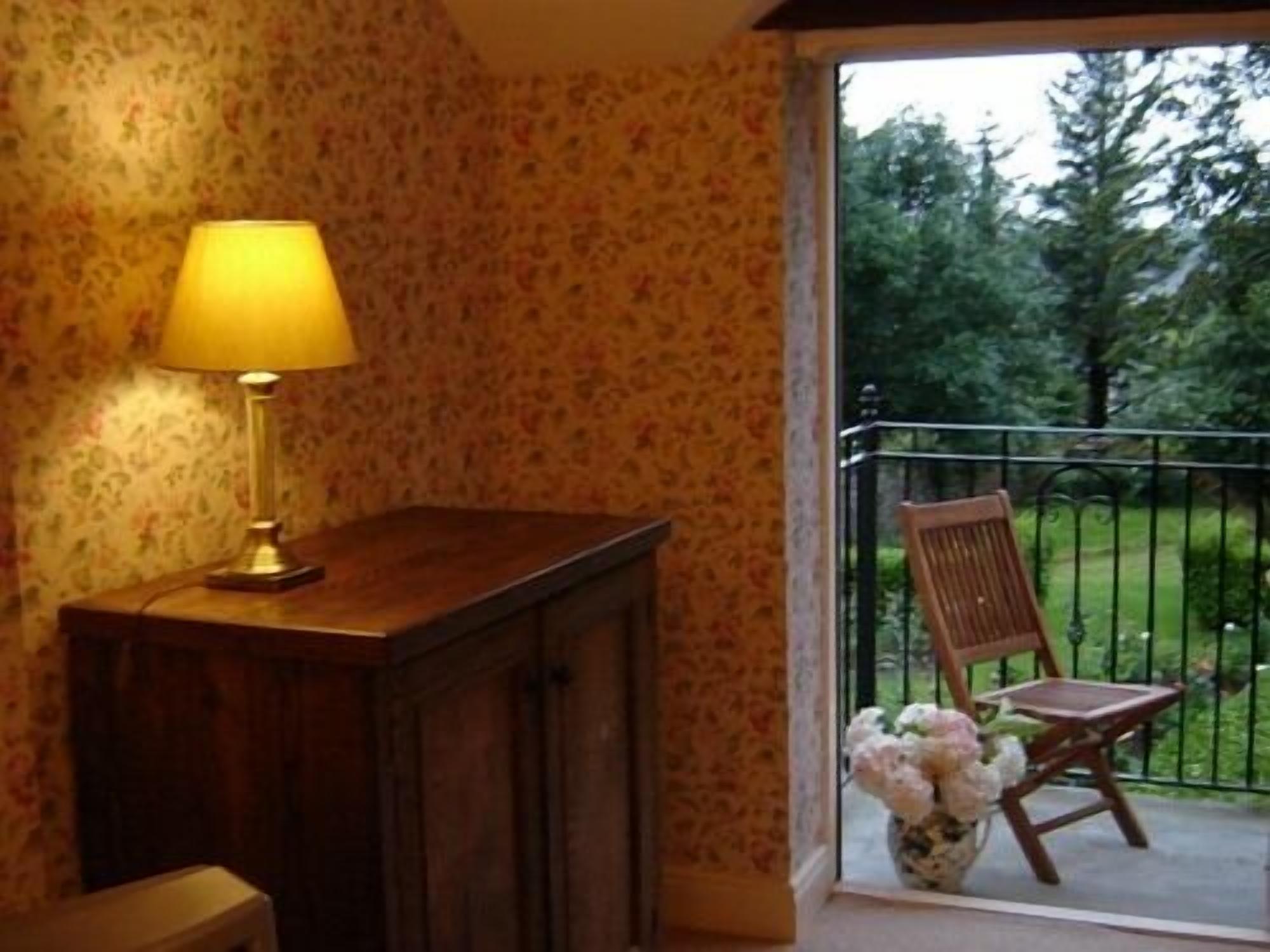 A room with floral wallpaper, a wooden cabinet, a table with a lamp, a chair, and a balcony with a view of trees.