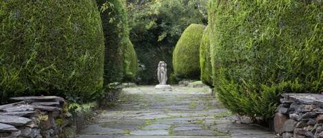 Un chemin de pierre bordé de buissons taillés en formes géométriques, qui mène à une statue.