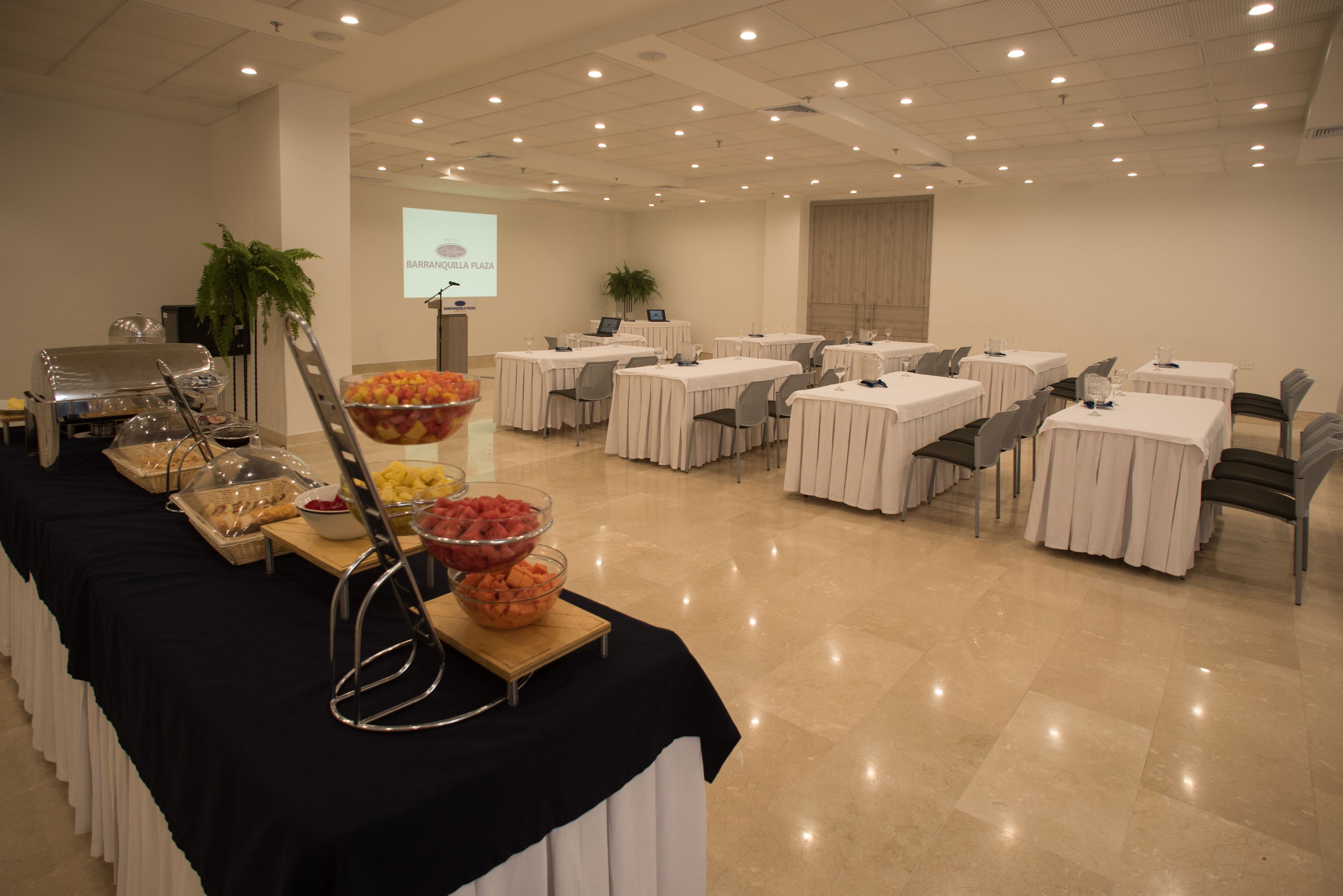 Un salón de conferencias con mesas dispuestas para una reunión, una pantalla de proyector y una mesa de buffet con diversas opciones de comida.