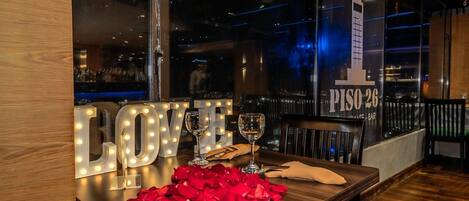 A table with illuminated 'LOVE' letters, two wine glasses, and scattered rose petals.