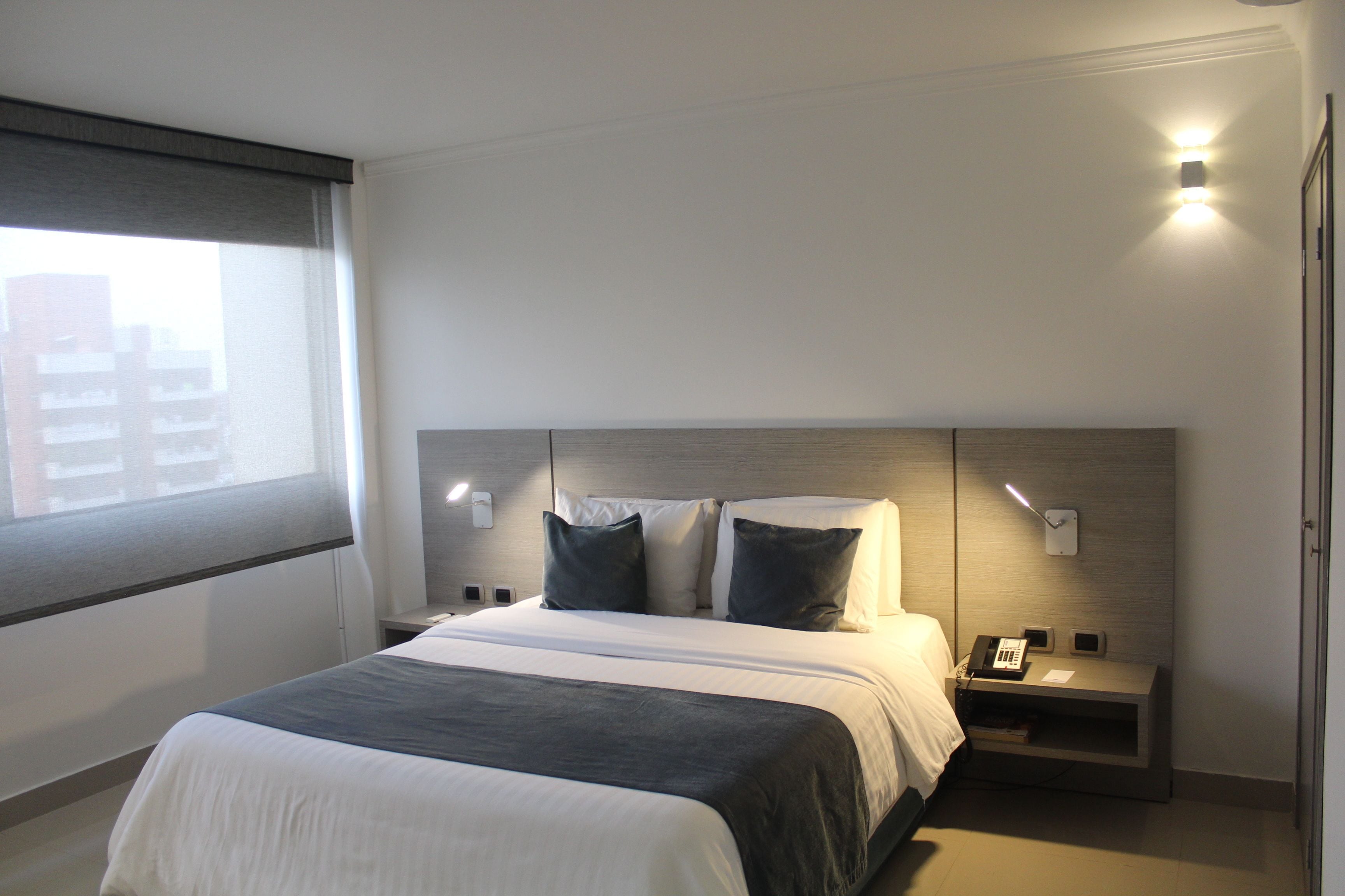 A modern hotel room with a large bed, bedside tables, a nightstand, and a window with blinds.
