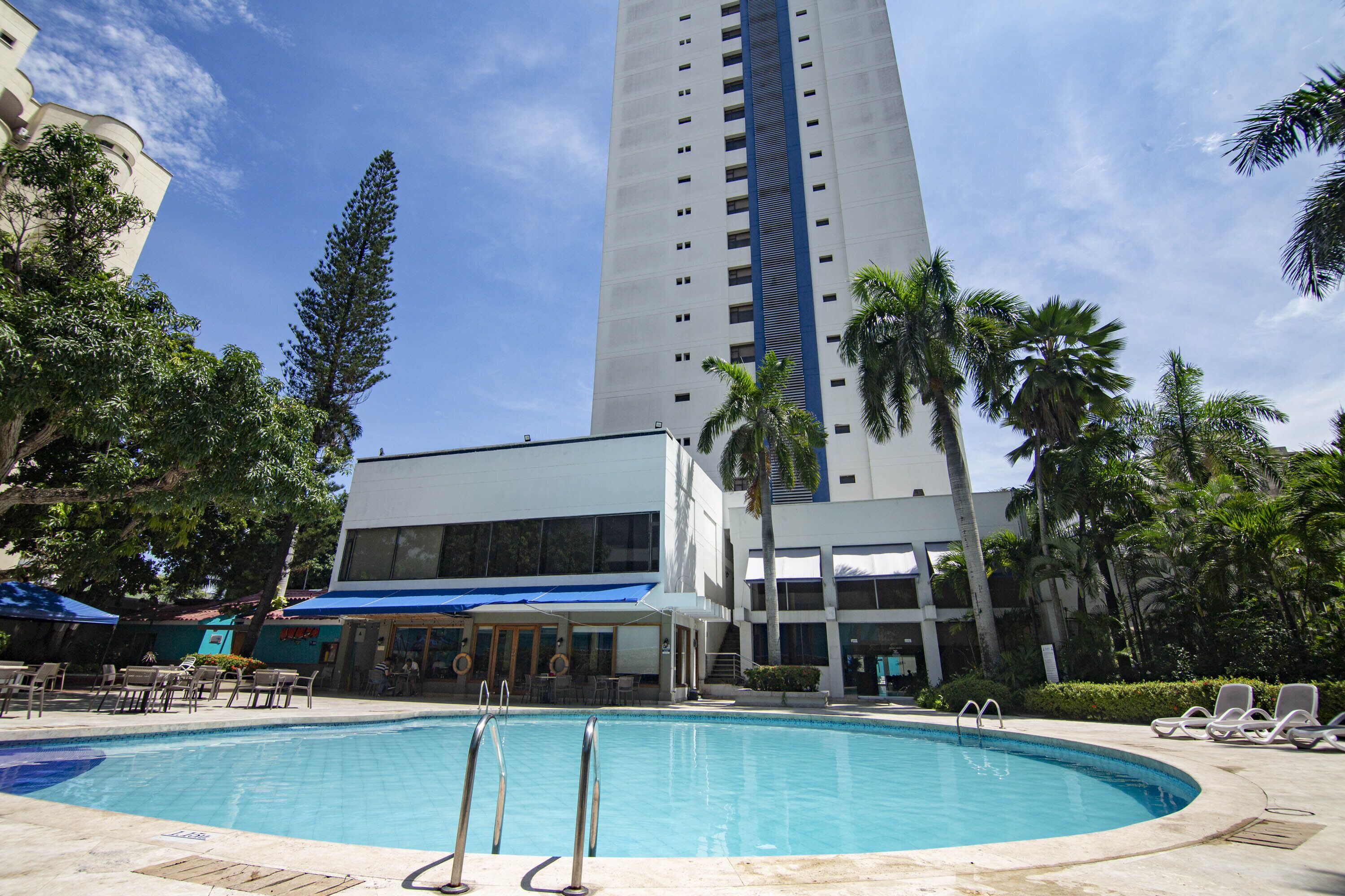 A swimming pool with lounge chairs, a tall building in the background, and palm trees.