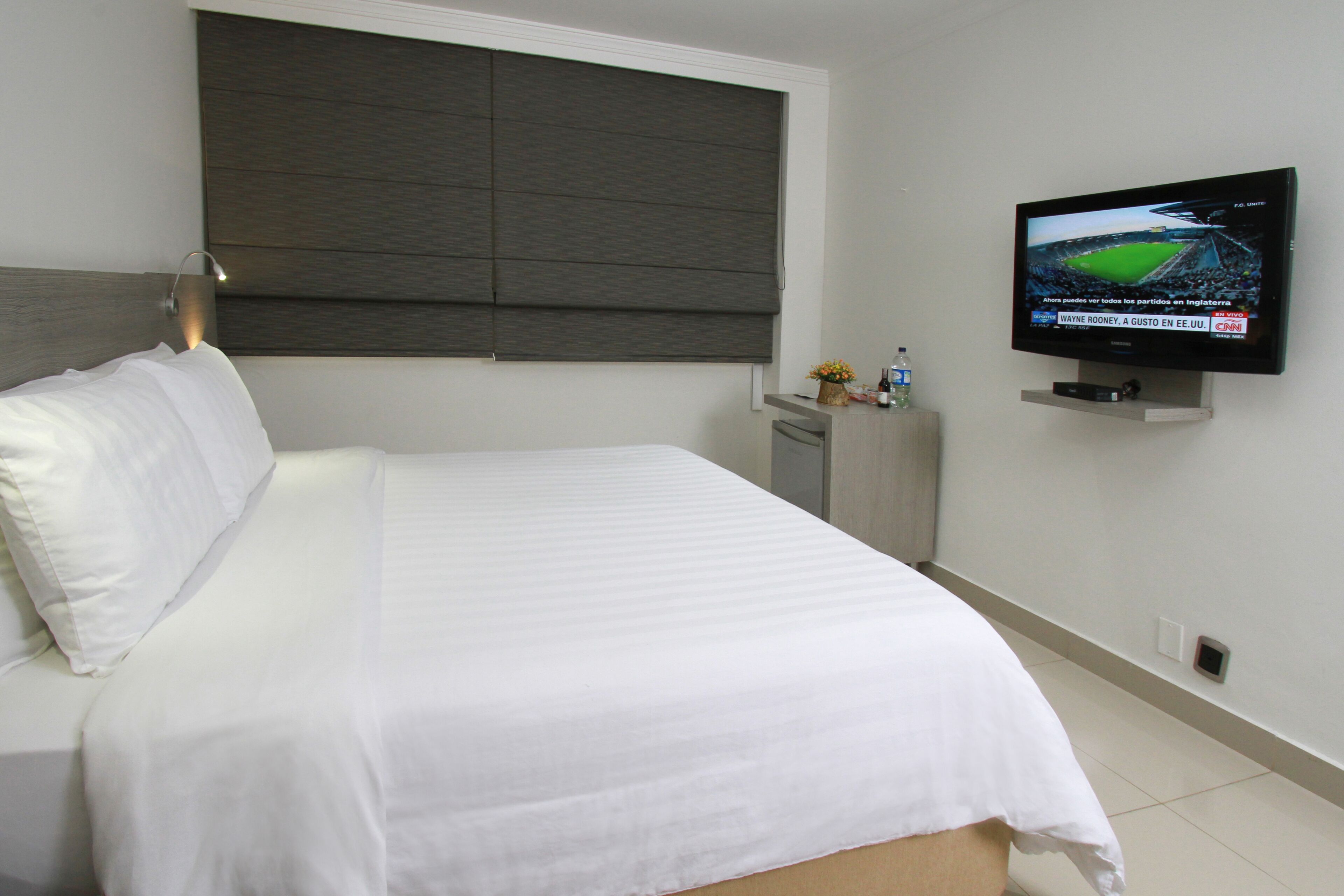 A hotel room with a bed, a television mounted on the wall, and a window with a dark blind.