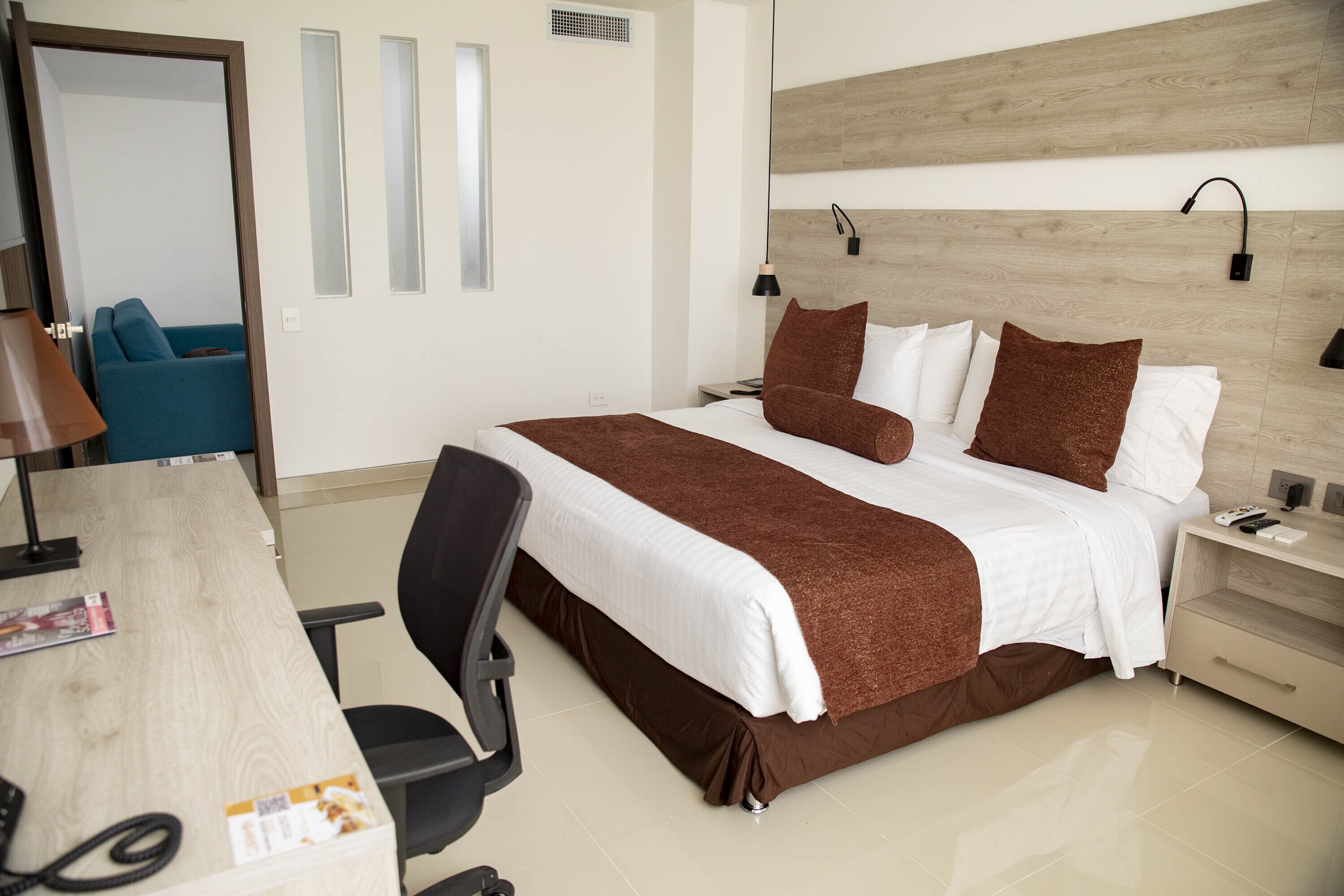 Una habitación de hotel con cama, mesita de noche, lámpara, sillón y escritorio con teléfono.
