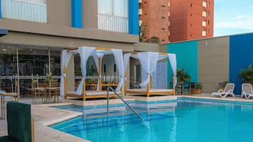 Zona de piscina de hotel con piscina azul cristalina, tumbonas, una zona de estar cubierta y vistas del edificio del hotel.
