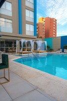 Zona de piscina de hotel con piscina azul cristalina, tumbonas, una zona de estar cubierta y vistas del edificio del hotel.