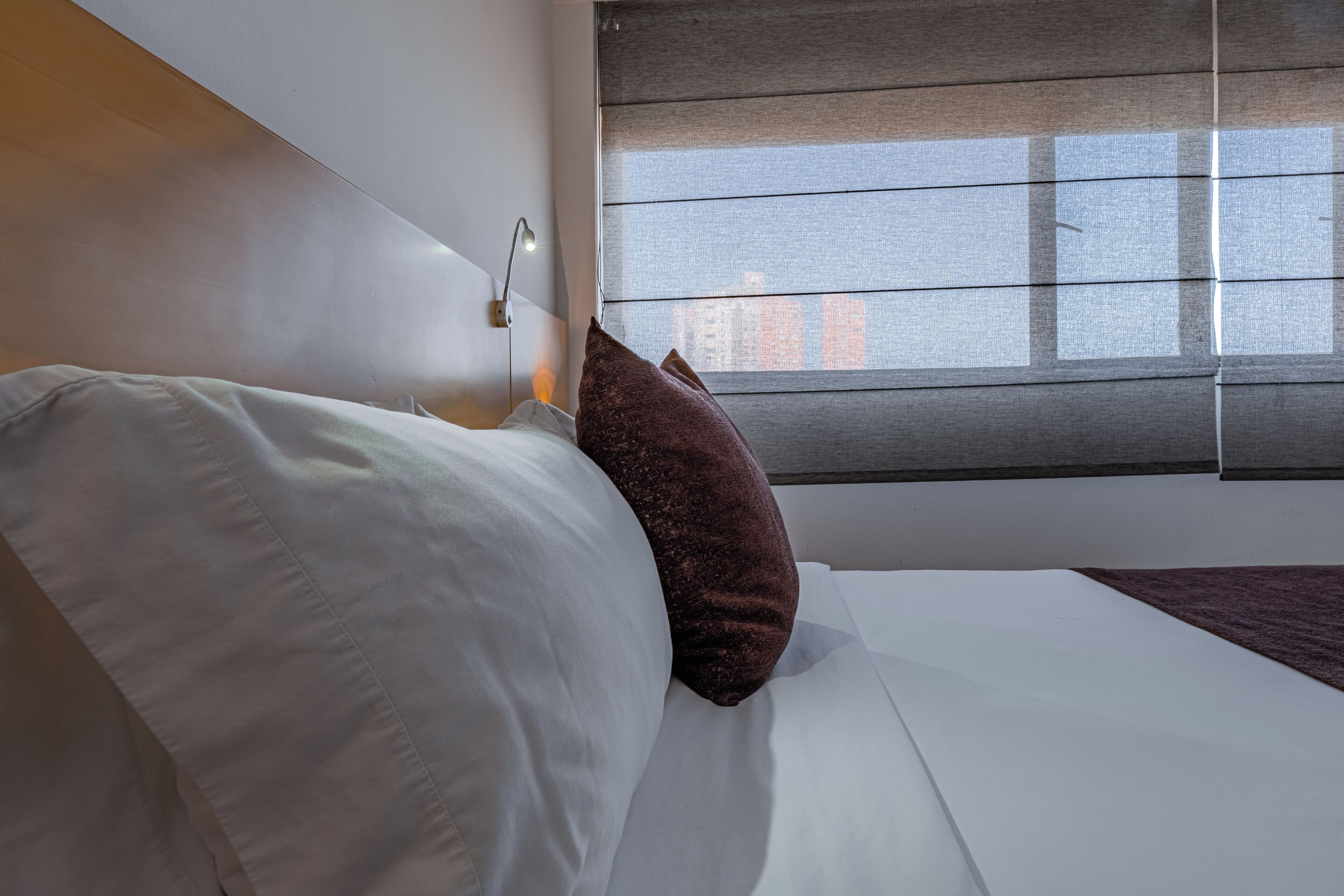 A hotel room with a bed, white bedding, a brown pillow, a nightstand with a lamp, and a window with blinds.