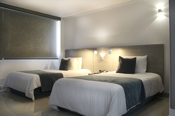 A hotel room with two beds, each with a headboard, white bedding, and a dark-colored throw blanket. There are wall-mounted lights above each bed, and a large window on the left side.