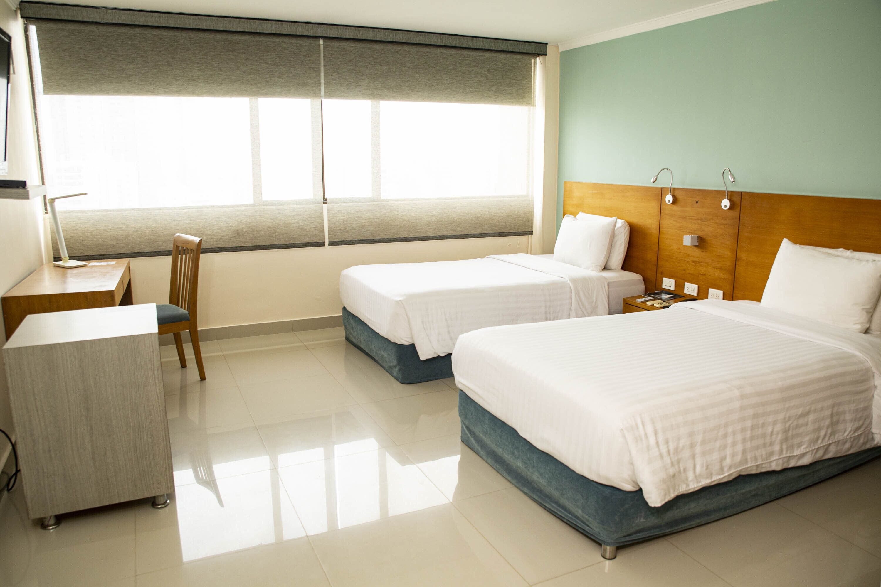 Habitación de hotel con dos camas individuales, un escritorio, una silla y un ventanal grande con persianas.