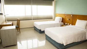 A hotel room with two single beds, a desk, a chair, and a large window with blinds.