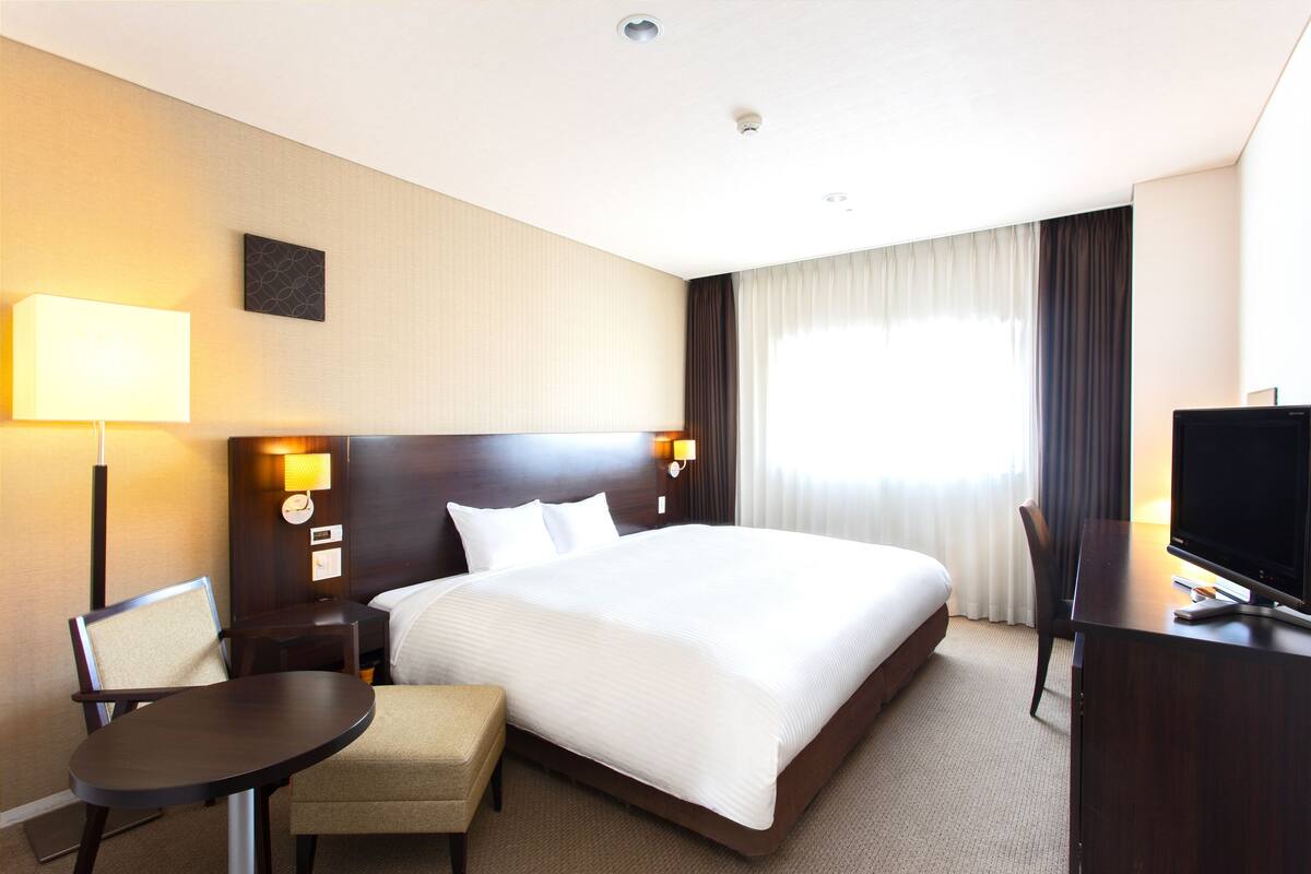 A hotel room with a large bed, a desk with a chair, a TV, a lamp, and a small table.