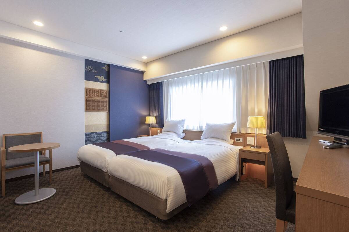 A hotel room with a large bed, a desk, a chair, a TV, and a window with curtains.