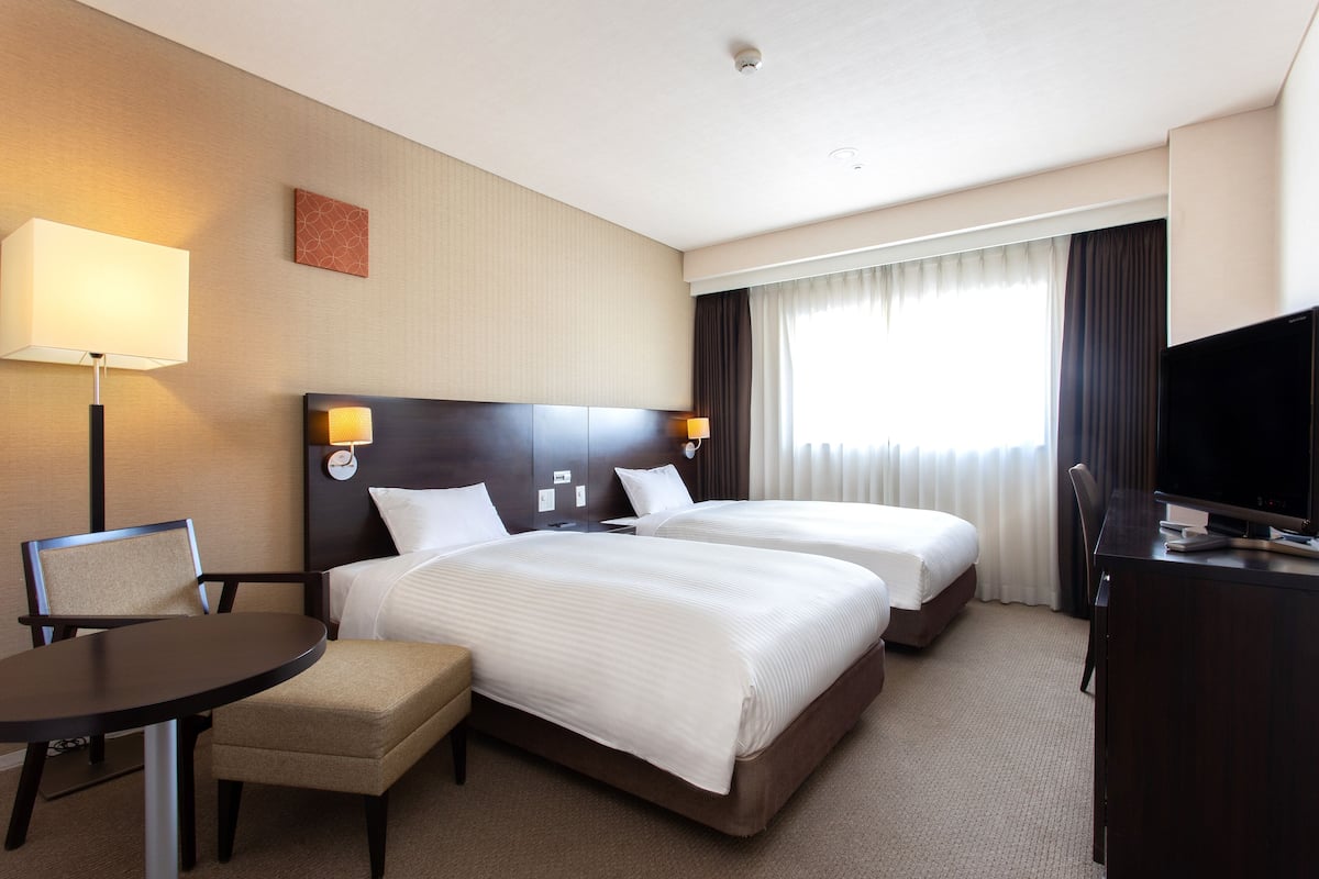 A hotel room with two beds, a desk, a chair, a TV, and a lamp.