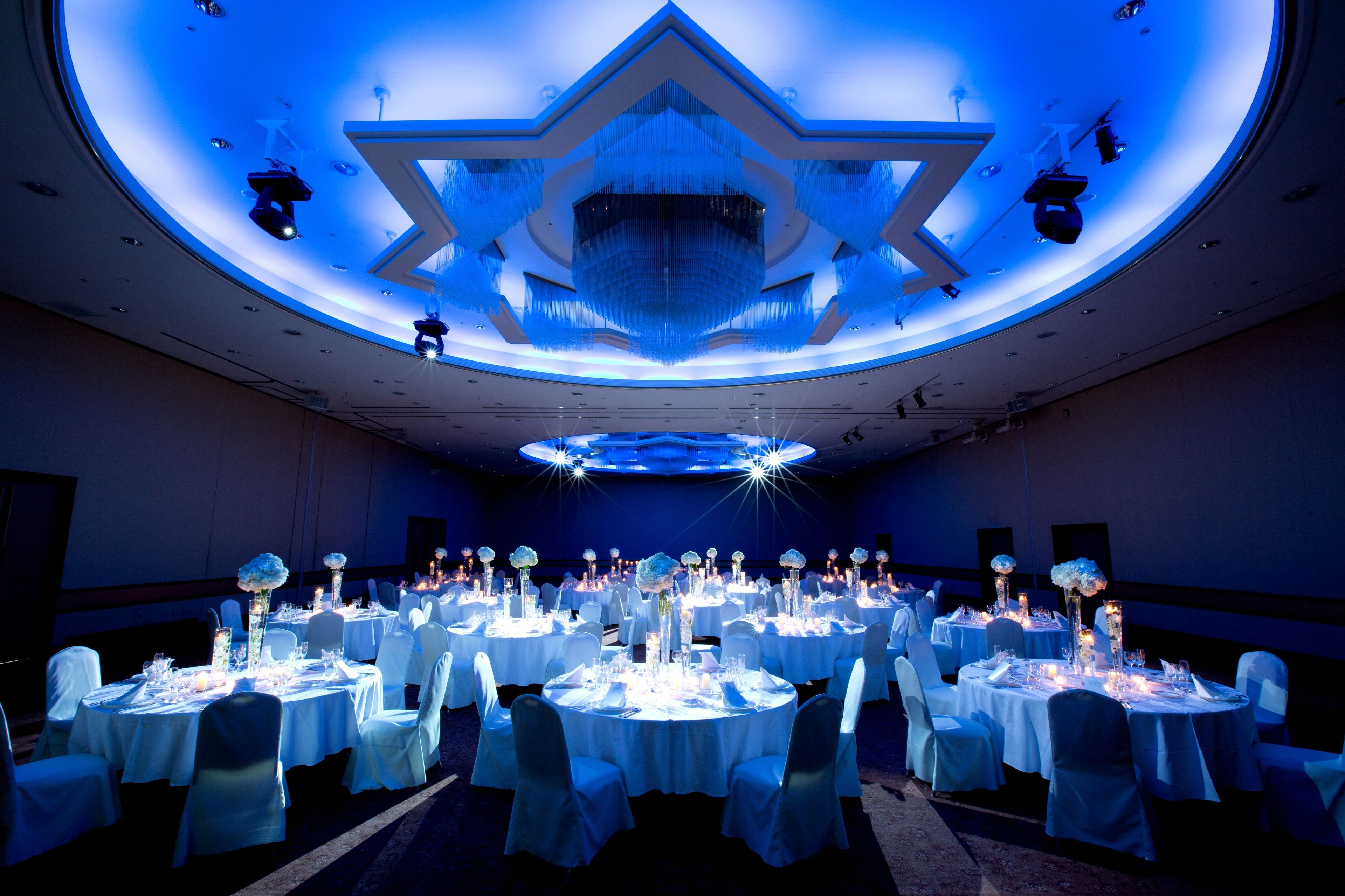 Un salón de banquetes con mesas redondas dispuestas para un evento formal, con una lámpara central de araña y un diseño de techo en forma de estrella.