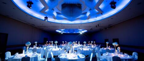 Un salón de banquetes con mesas redondas dispuestas para un evento formal, con una lámpara central de araña y un diseño de techo en forma de estrella.