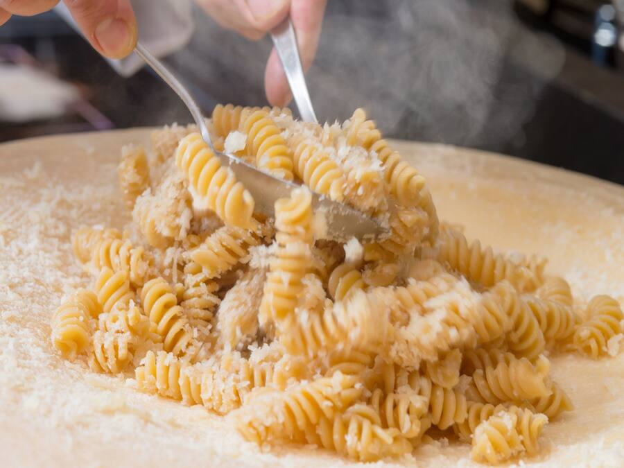Nahaufnahme einer Person, die mit einem Löffel Pasta mit geriebenem Käse auf einer flachen Oberfläche vermischt.