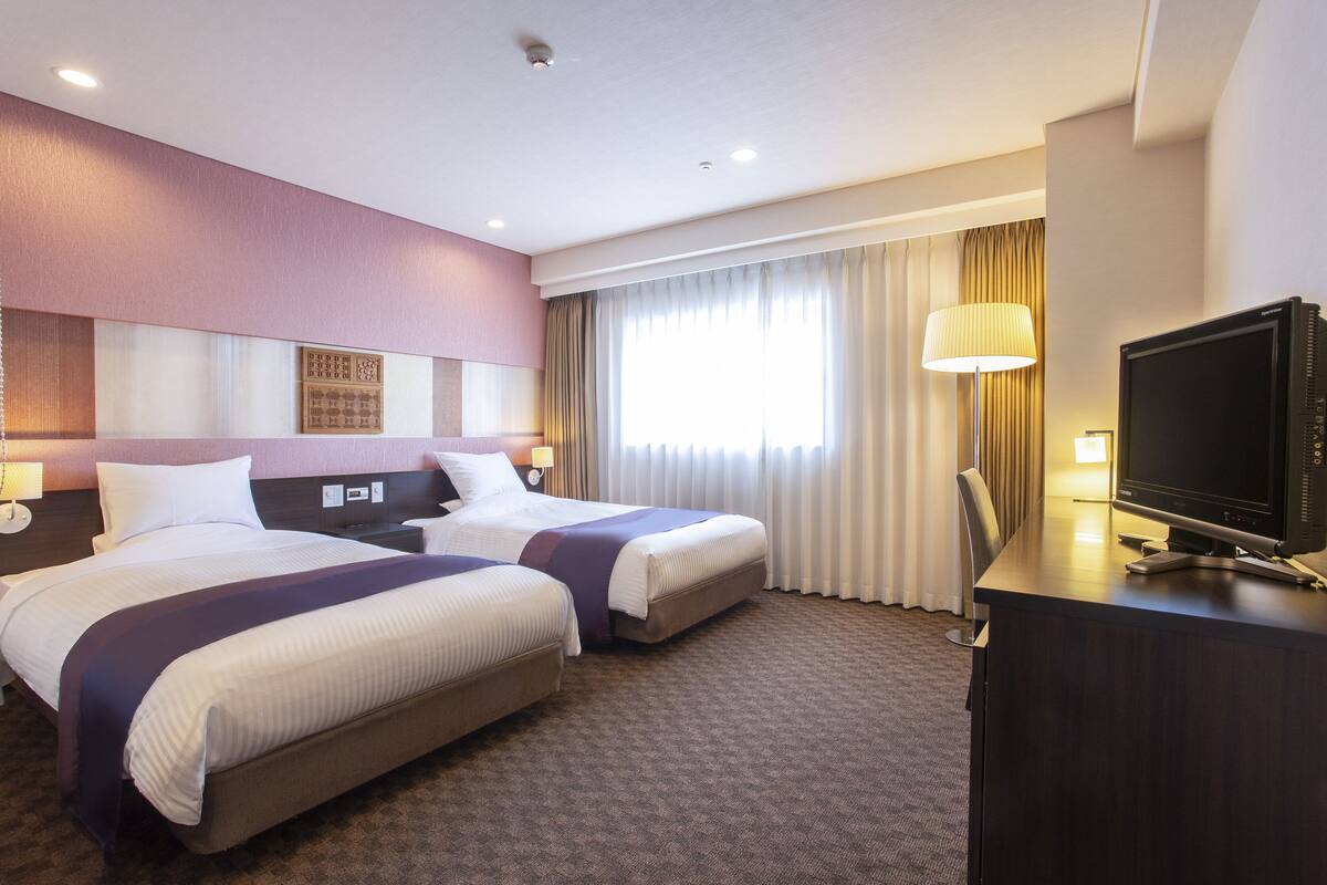 A hotel room with two beds, a desk with a TV, and a window with curtains.