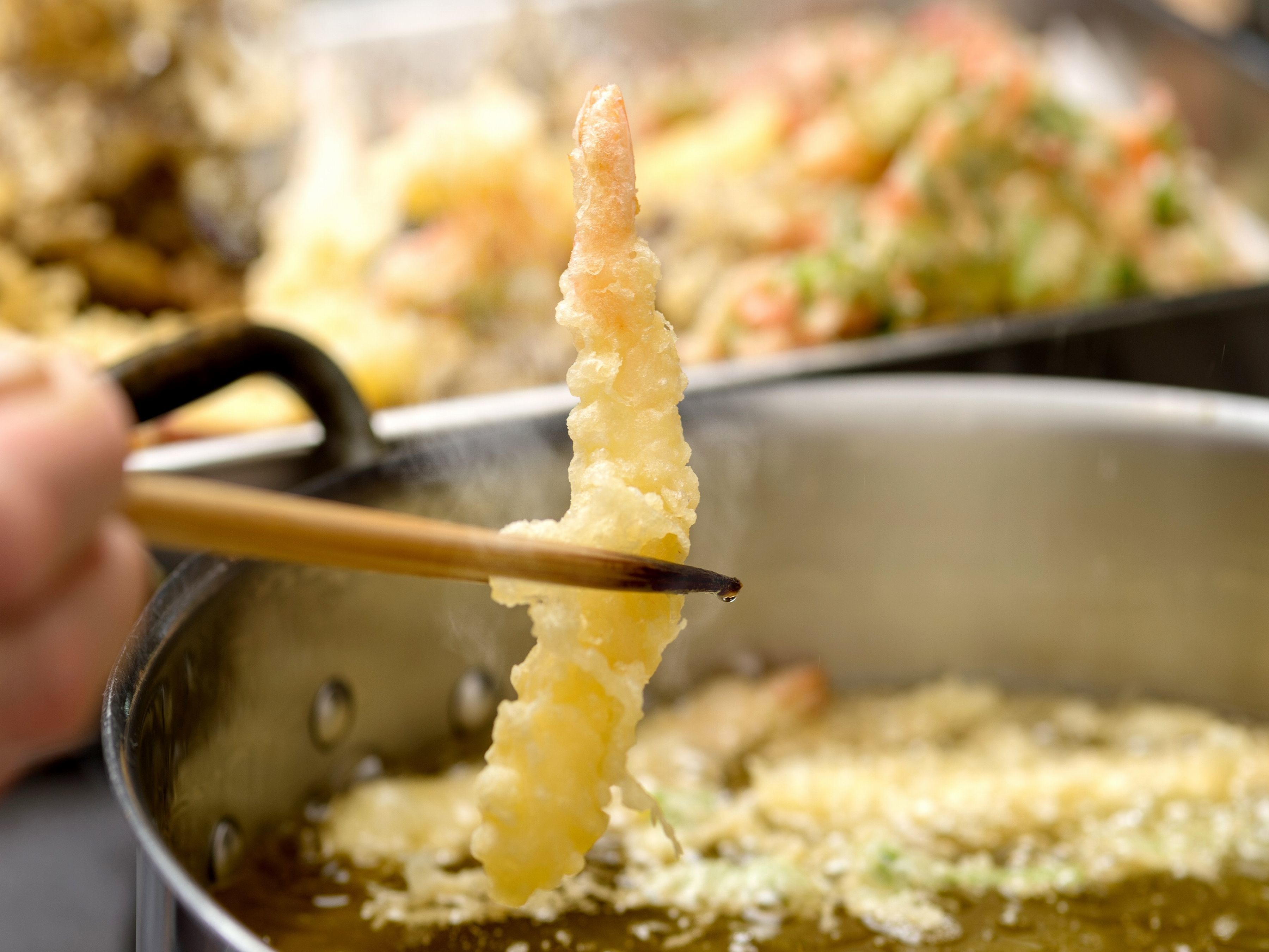 A close-up of a hand holding a piece of tempura with chopsticks over a pan of oil.