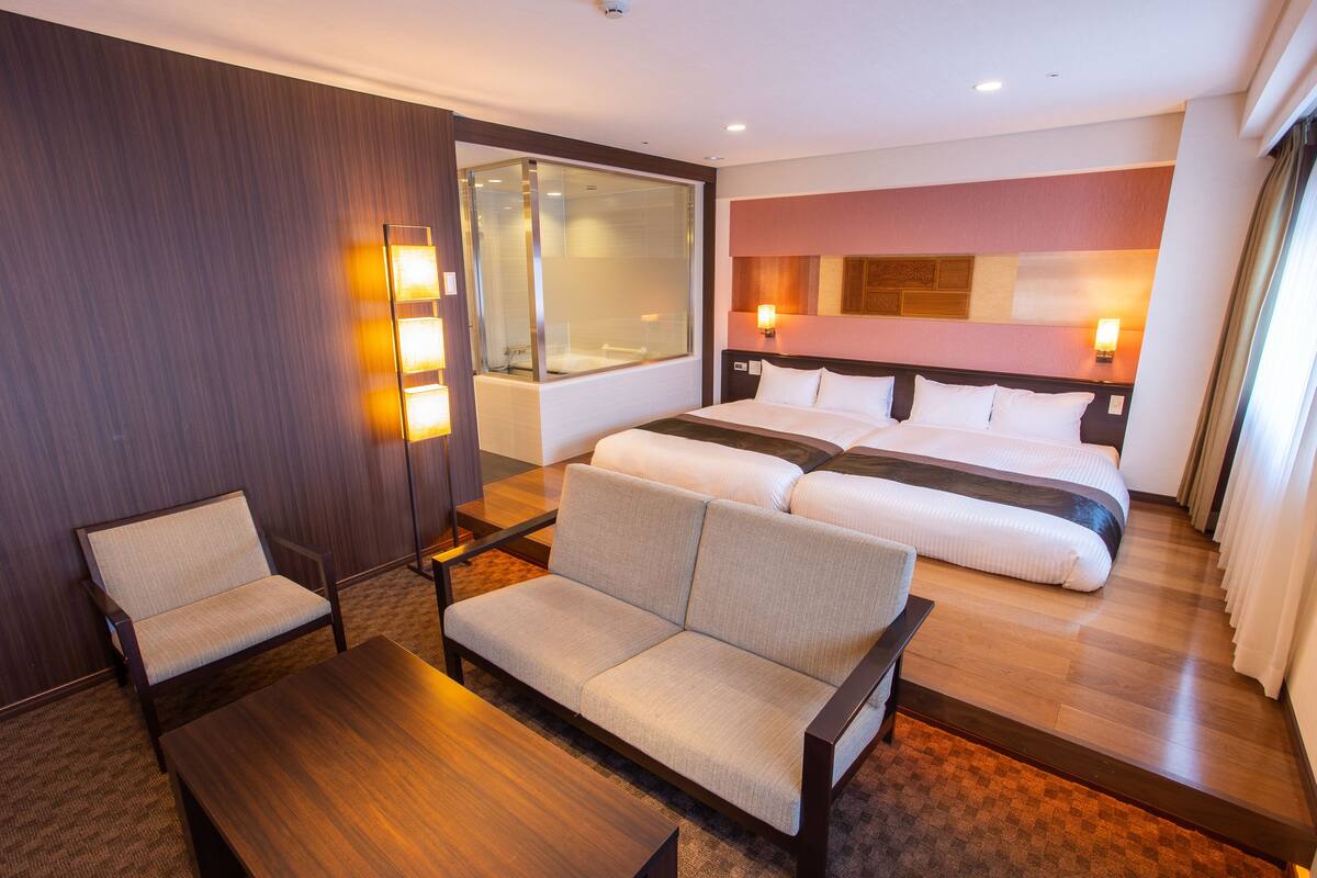 A hotel room with a large bed, a sofa, a chair, and a desk. There is a window with curtains and a wall-mounted light fixture.