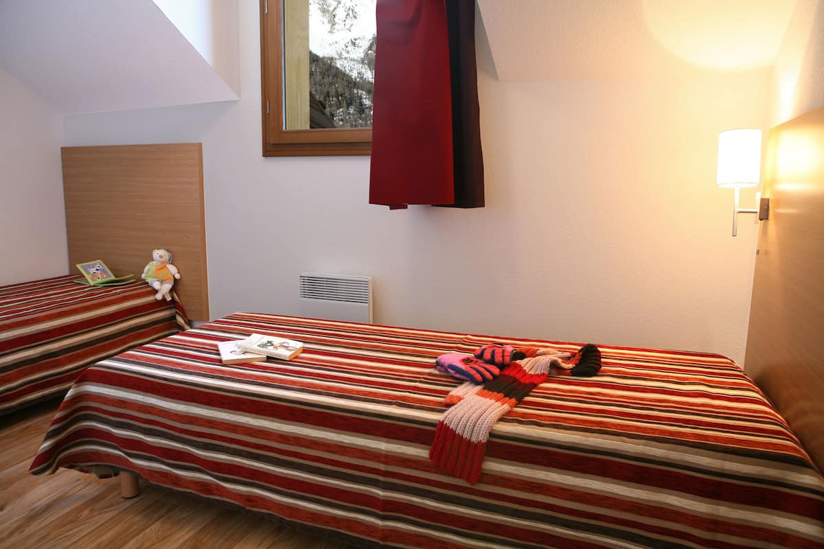 Une chambre avec deux lits, chacun agrémenté de draps à rayures, une fenêtre donnant sur un paysage agréable, une lampe fixée au mur et un rideau rouge.