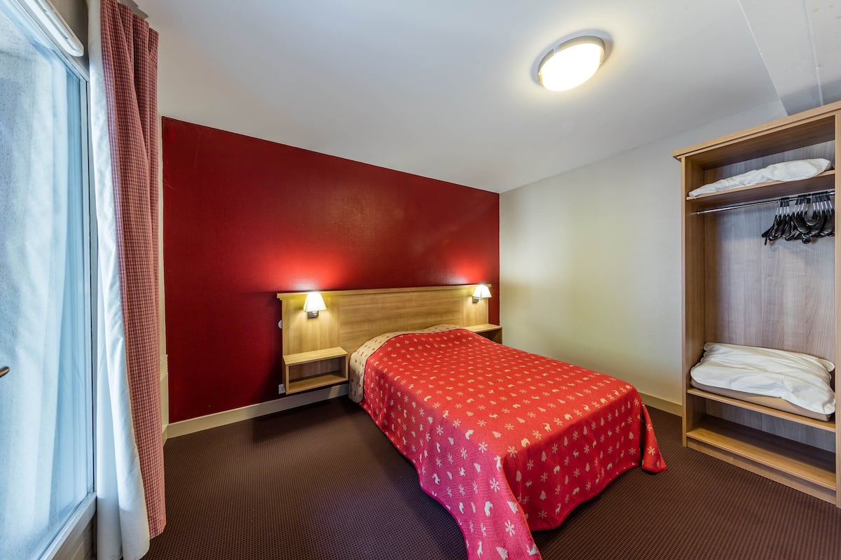 Une chambre d’hôtel avec un mur rouge, une tête de lit en bois, un lit simple avec une couverture rouge, une armoire où sont suspendus des vêtements et une table de chevet avec une lampe.
