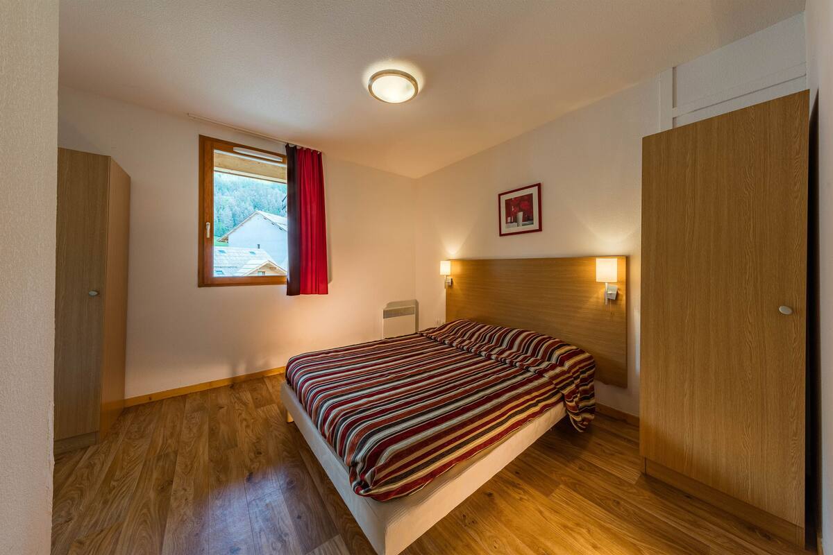 Une chambre avec un plancher en bois, un lit avec une literie à rayures, une fenêtre avec des rideaux rouges, une lampe de chevet et un tableau encadré au mur.