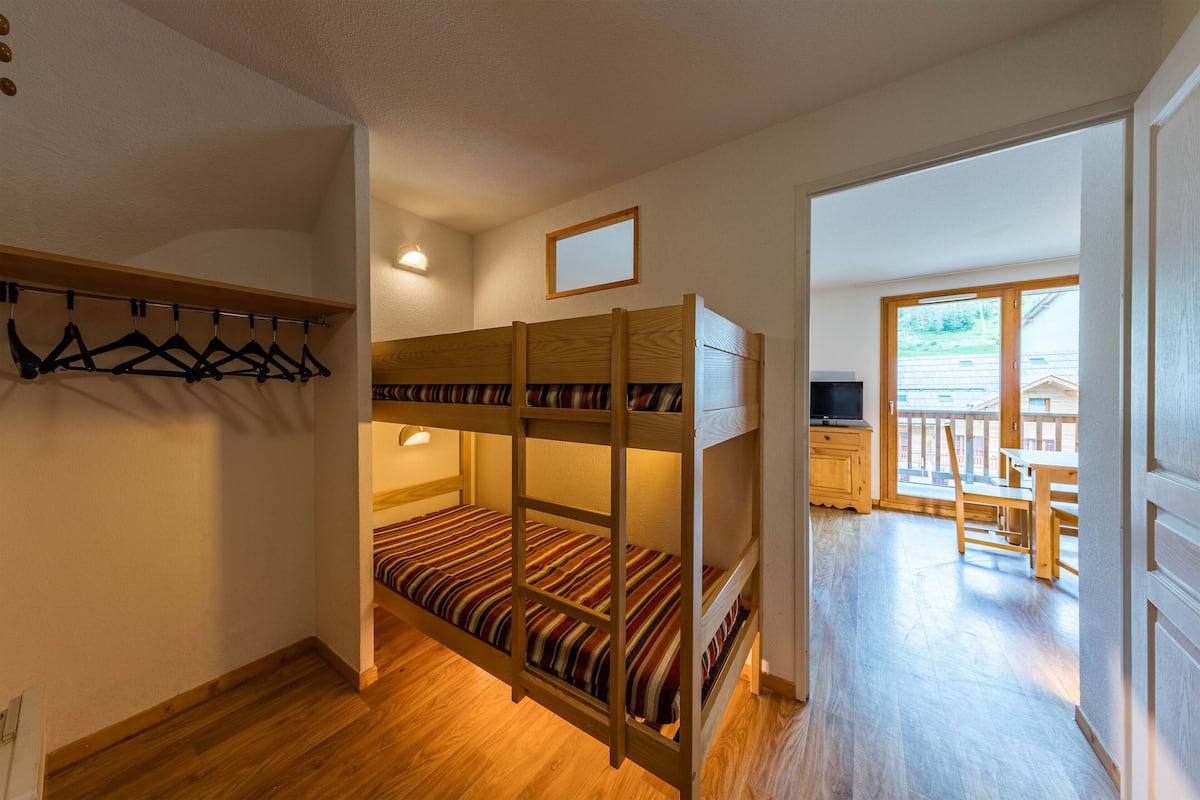 Un lit superposé avec une literie à rayures, un bureau en bois, une chaise et un téléviseur dans une pièce dotée de planchers en bois et d’un balcon.