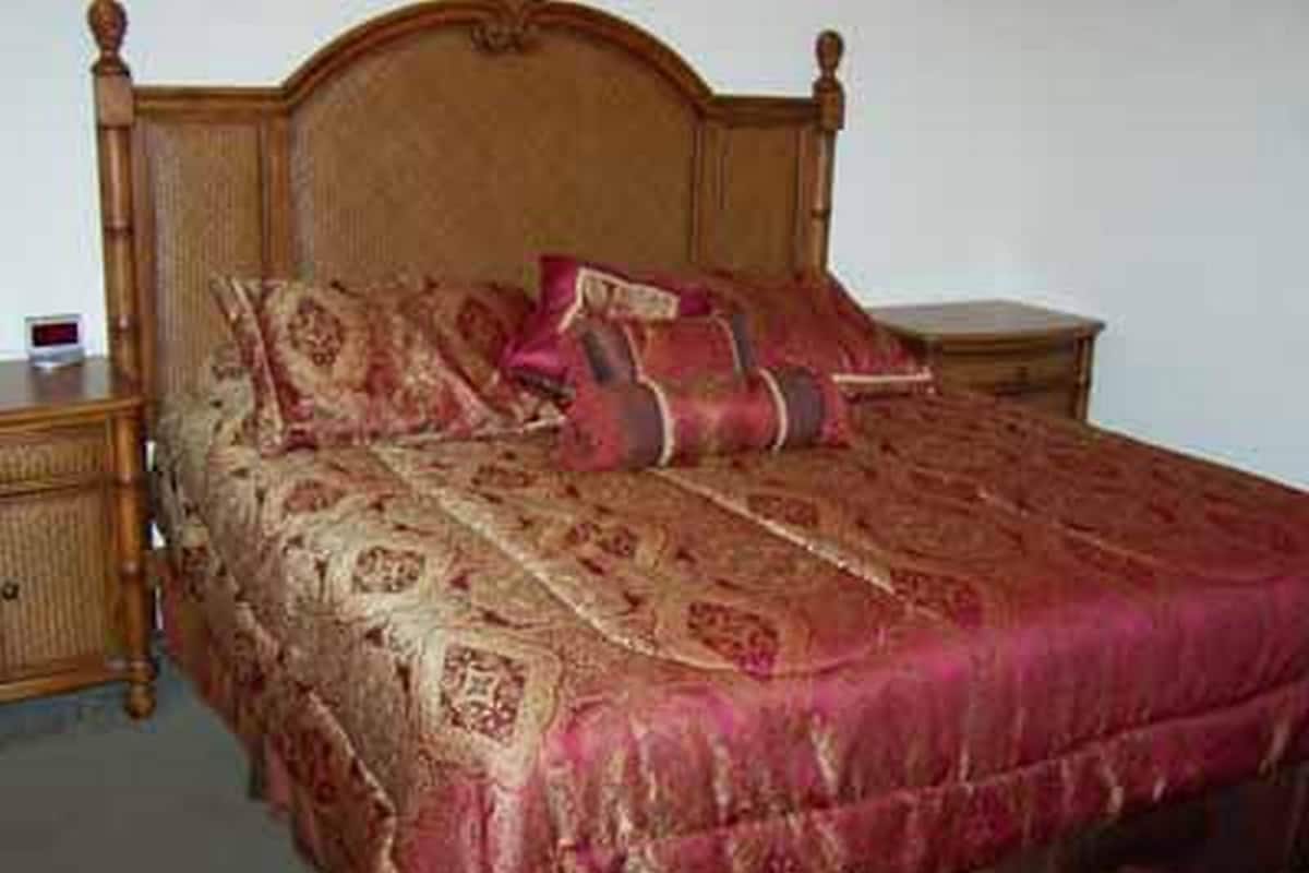Ein hölzernes Bett mit Kopfstück, einer rot-goldenen Tagesdecke und zwei Kissen.
