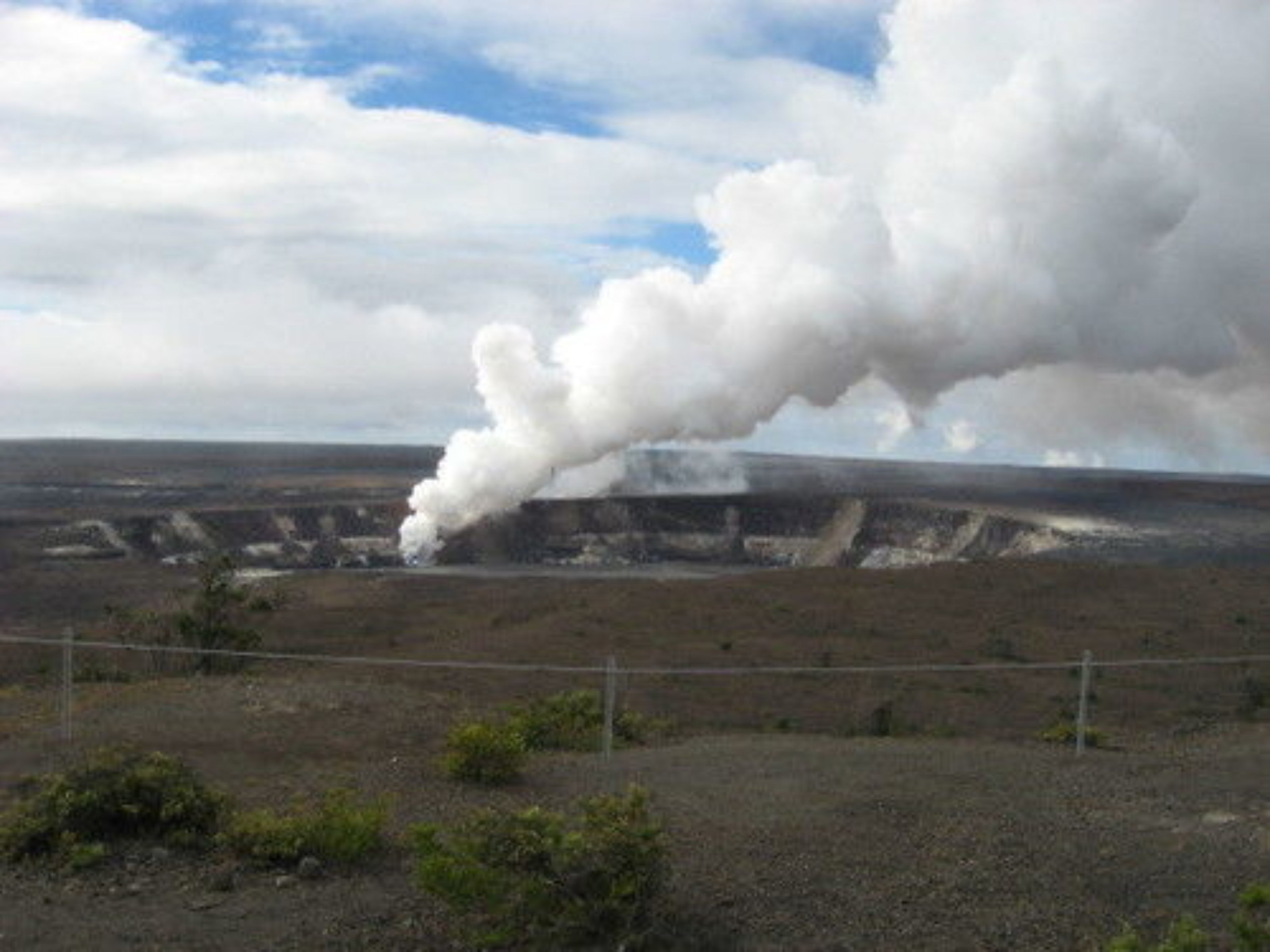 A barren landscape with a large plume of smoke rising from a distant structure.
