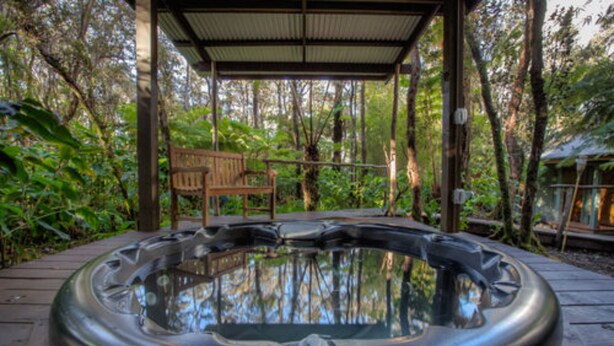 A hot tub with a wooden deck and a wooden bench under a covered area surrounded by trees.