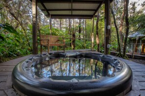 A hot tub with a wooden deck and a wooden bench under a covered area surrounded by trees.