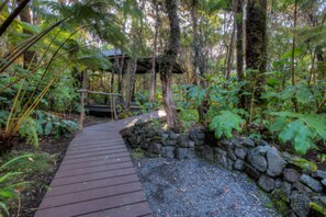 A wooden pathway leading through a lush garden with a stone wall and a wooden structure in the background.