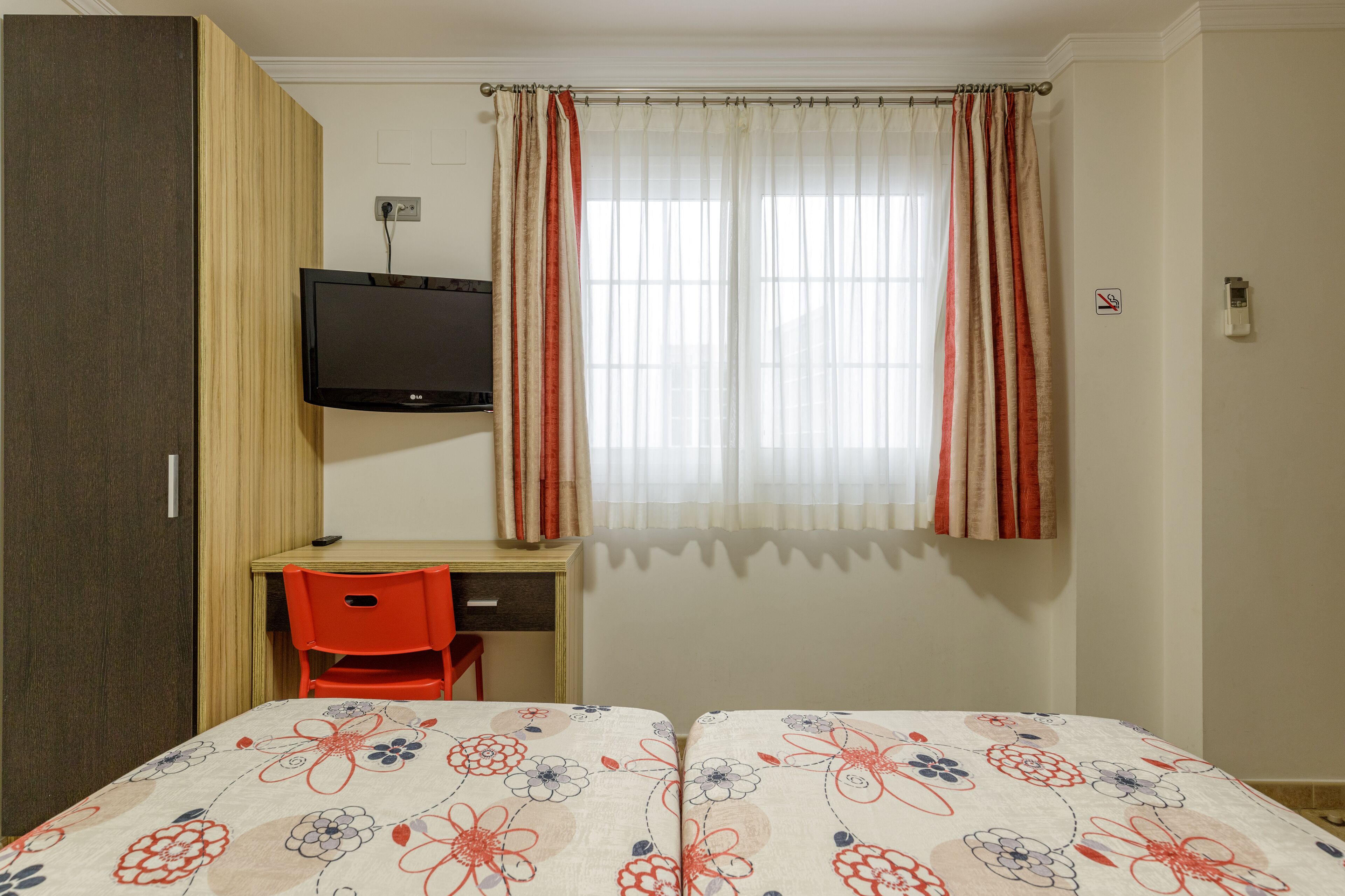 Una habitación con cama, un escritorio con silla, televisor y ventana con cortinas.