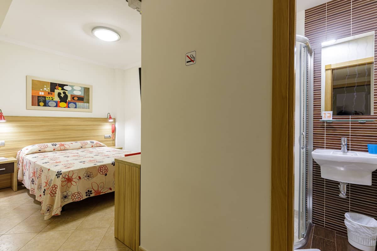 A hotel room with a bed, a nightstand, a wall-mounted painting, a bathroom with a sink and mirror, and a trash bin.