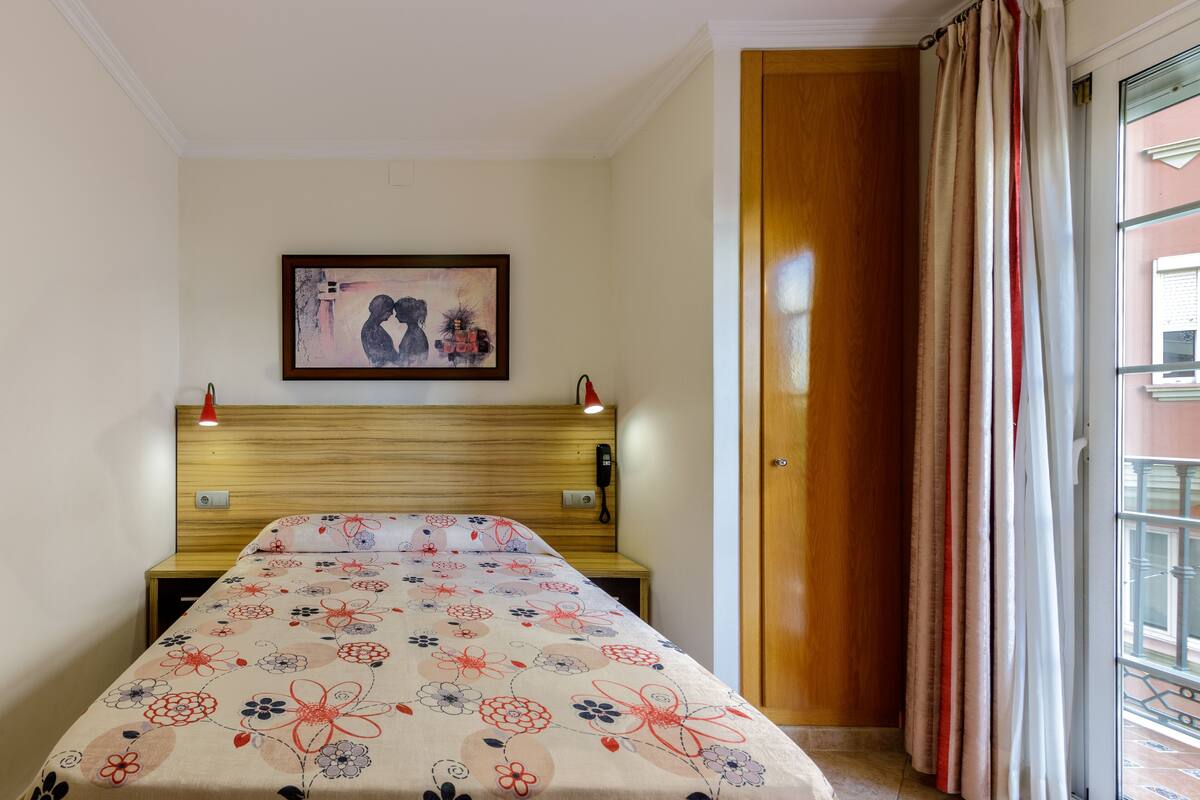 A room with a bed, a wooden headboard, a painting on the wall, a telephone, and a window with curtains.