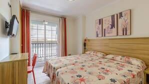 A hotel room with a wooden desk, two single beds with floral bedding, a TV mounted on the wall, and a balcony with red curtains. -  HA Hotel Trebol (Málaga)