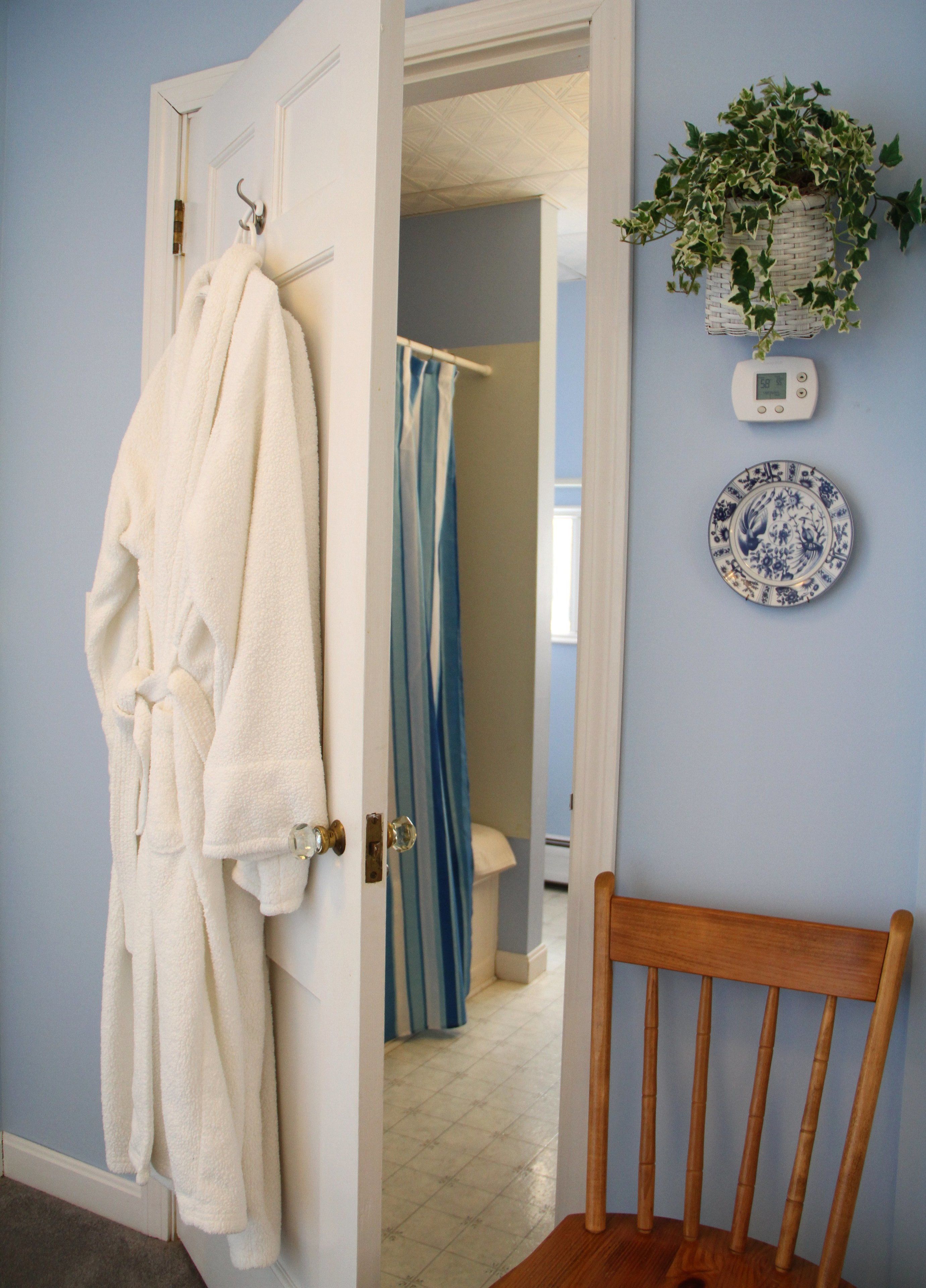 A bathroom with a blue shower curtain, a white bathrobe hanging on a door, a wooden chair, and a wall-mounted scale.