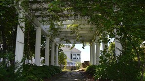 A covered stone pathway leading to a white building with columns, surrounded by lush greenery. - Brewster Inn (Dexter)
