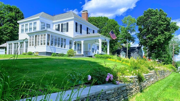 A large, white two-story house with a porch and a stone wall in front.