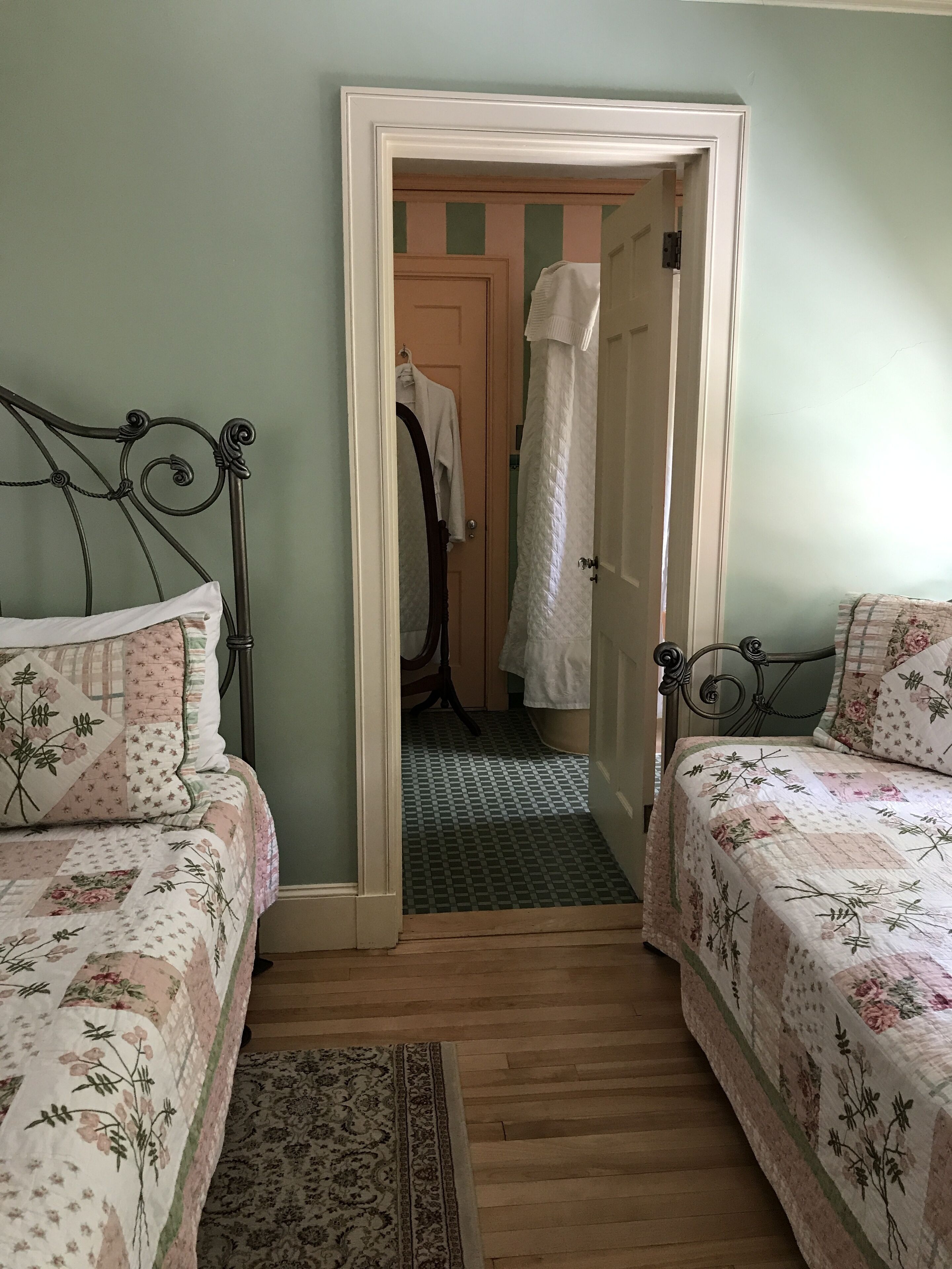 A bedroom with two beds, a wooden floor, a patterned rug, and a door leading to a bathroom.
