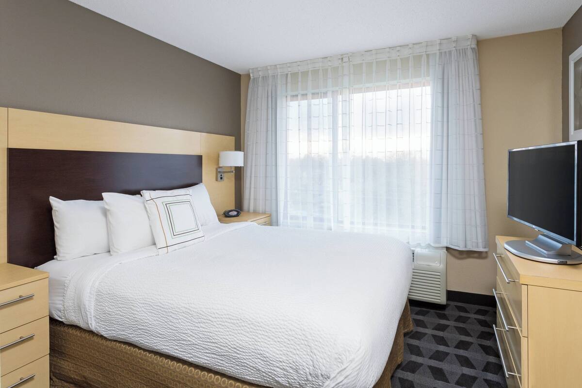 A hotel room with a large bed, a flat-screen TV, a bedside table, and a window with sheer curtains.
